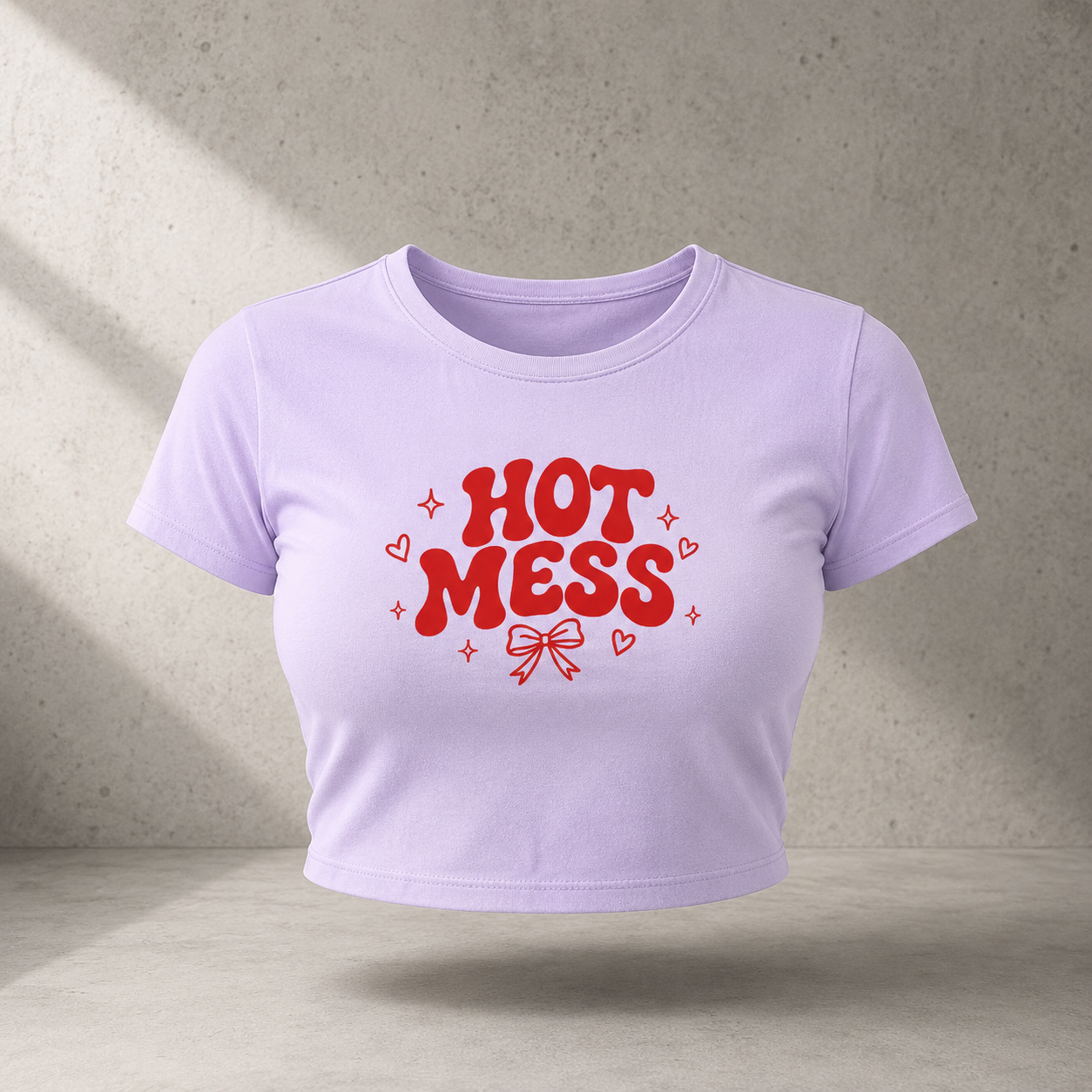 A light purple cropped short-sleeve t-shirt with a red graphic reading 'HOT MESS' in bubble letters, surrounded by small hearts and sparkles, shown against a textured concrete wall background.