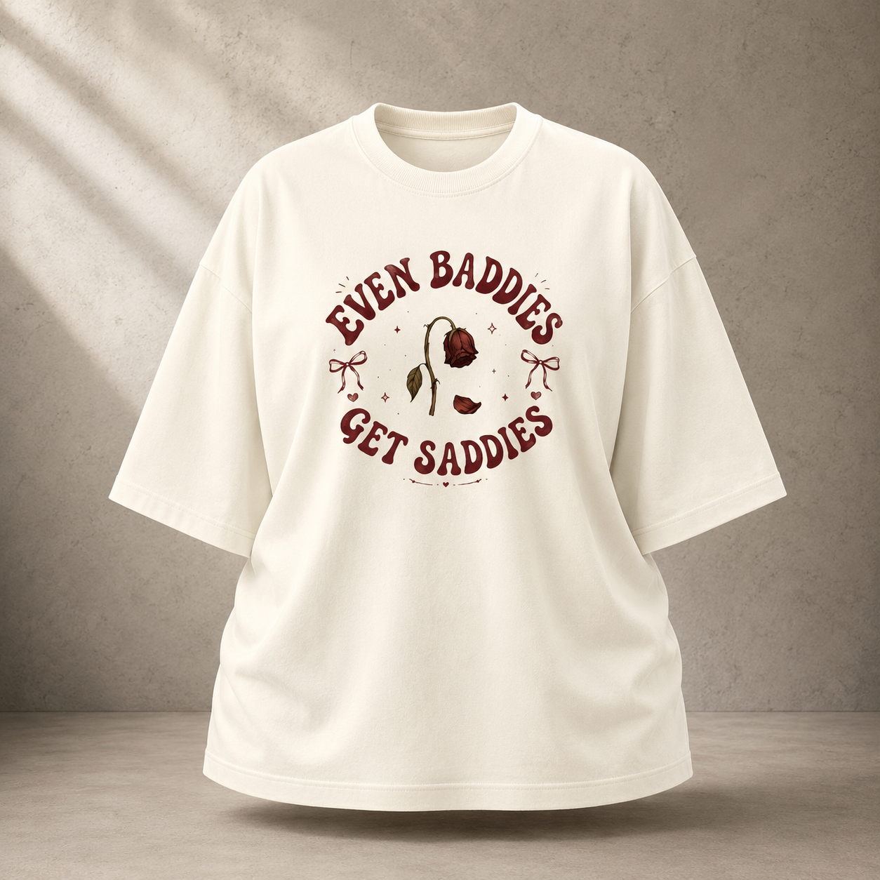 An oversized off-white t-shirt with a vintage-style graphic on the chest that reads 'EVEN BADDIES GET SADDIES' around a single rose illustration, displayed against a textured gray concrete wall background.