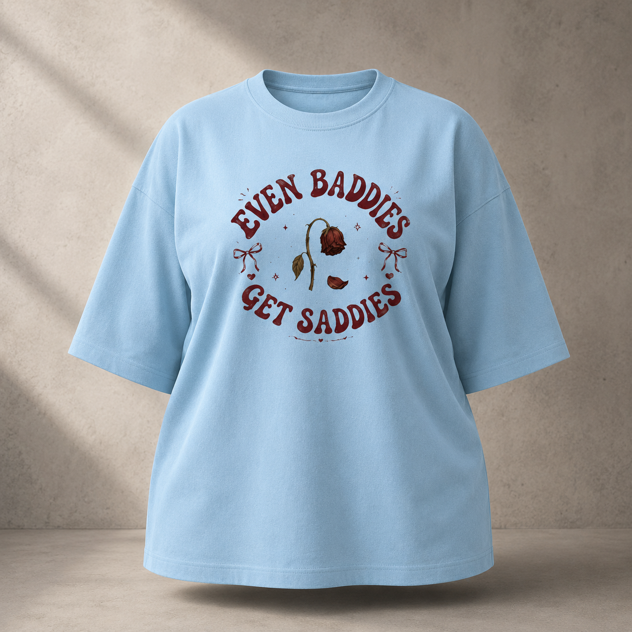 A babyblue oversized short-sleeve t-shirt with a vintage-style graphic on the chest reading 'EVEN BADDIES GET SADDIES' in dark red, surrounding a small illustration of a sad rose and a horse bit. The shirt is displayed against a textured beige wall with soft diagonal shadows.