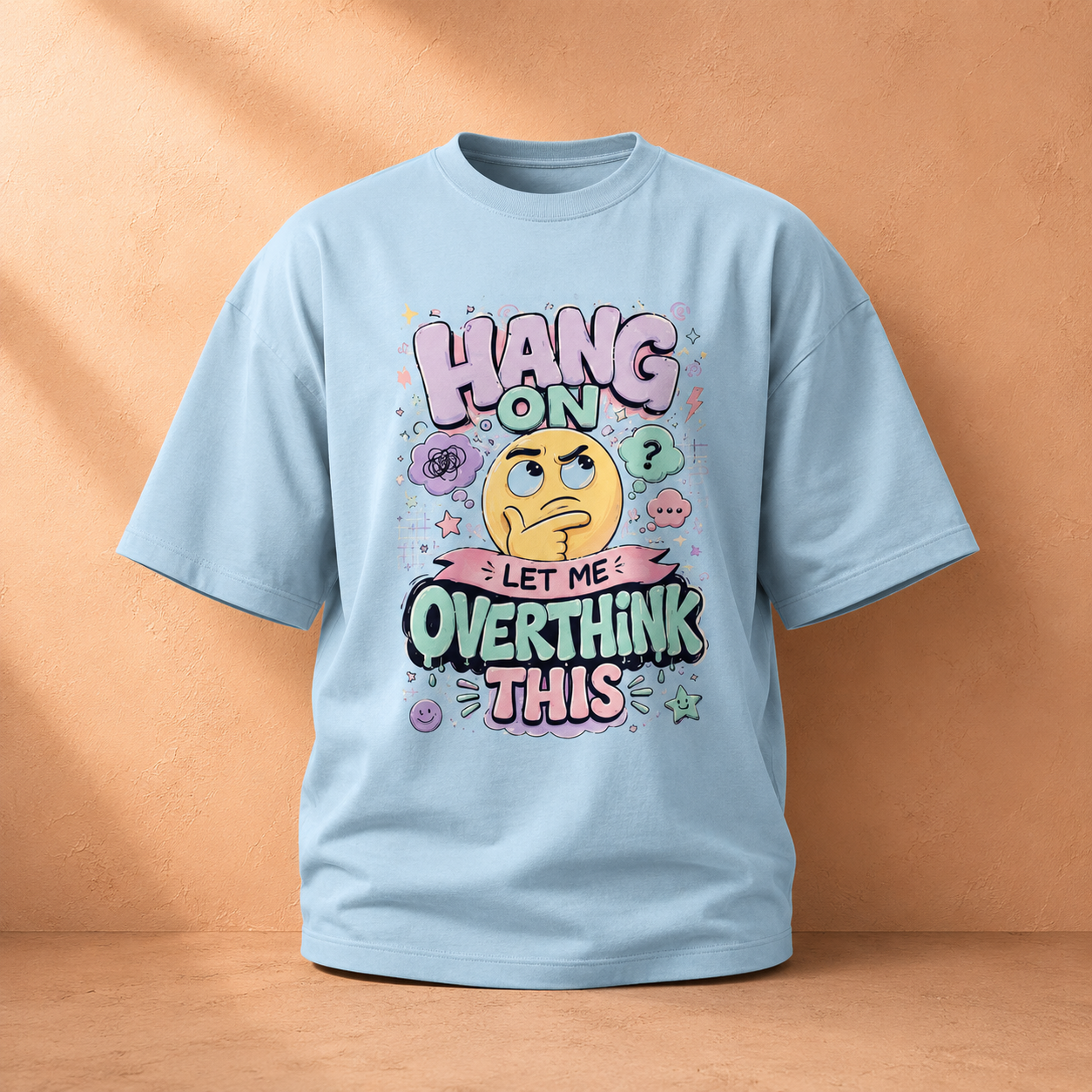 A light blue short-sleeve t-shirt with a colorful graphic on the front that reads 'HANG ON LET ME OVERTHINK THIS' around a thinking emoji, shown against a warm peach-colored background.