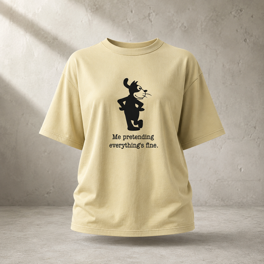 A light beige short-sleeve t-shirt with a black graphic of a cartoon cat and the text 'Me pretending everything's fine.' printed on the front, displayed against a textured gray wall.