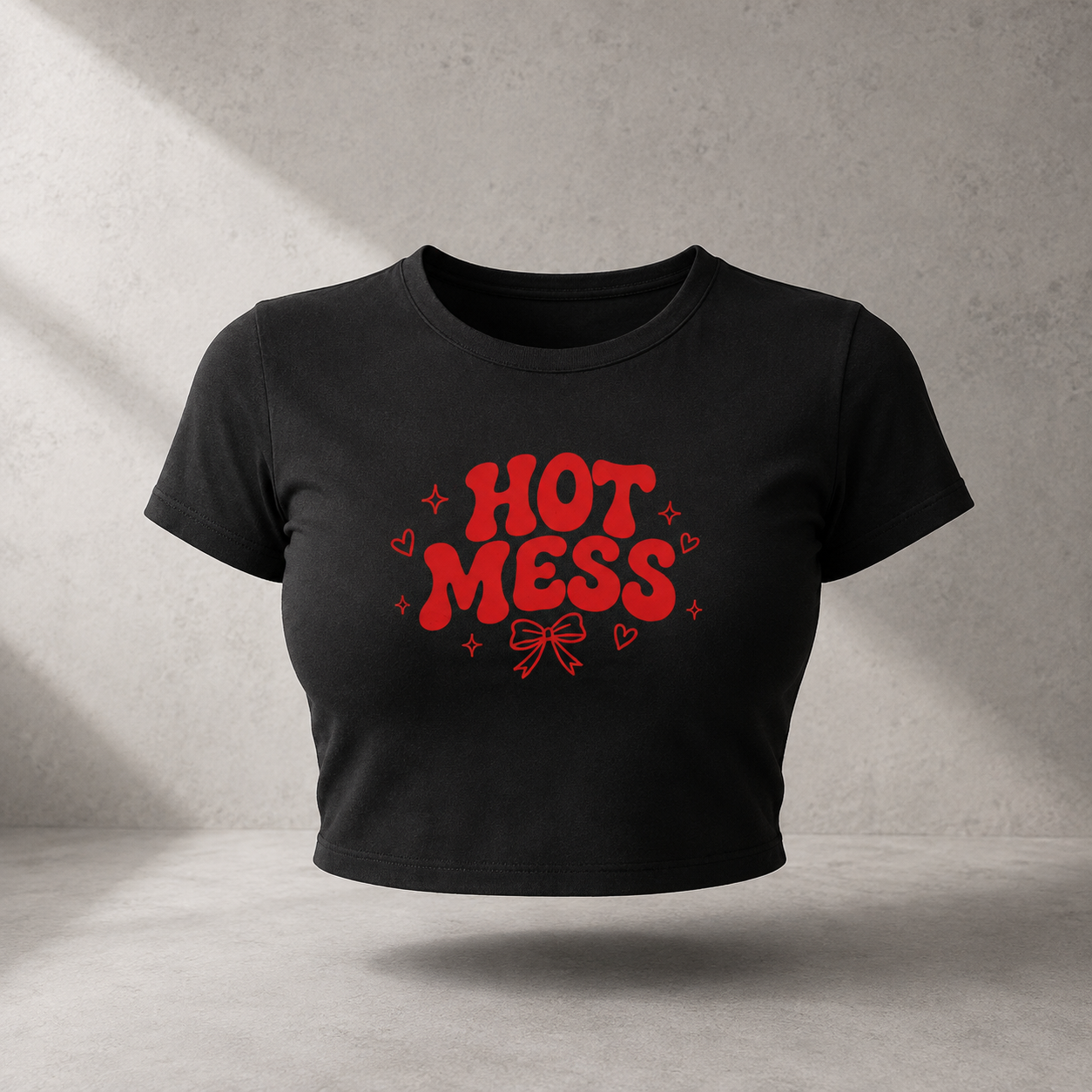 A black cropped short-sleeve t-shirt with a red graphic reading 'HOT MESS' in a bold retro font, surrounded by small hearts and stars, shown floating against a textured gray wall with soft diagonal shadows.