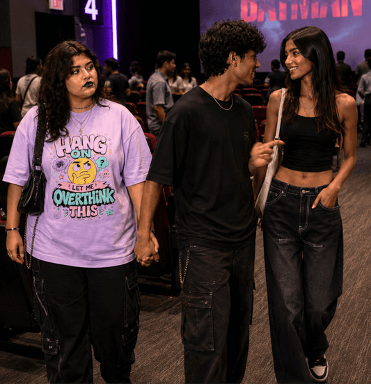 Three young adults walking and talking in a dimly lit event venue or backstage area. The person on the left wears a Lavender graphic T-shirt with a cartoon face and the text 'HANG ON LET ME OVERTHINK THIS,' paired with black pants.