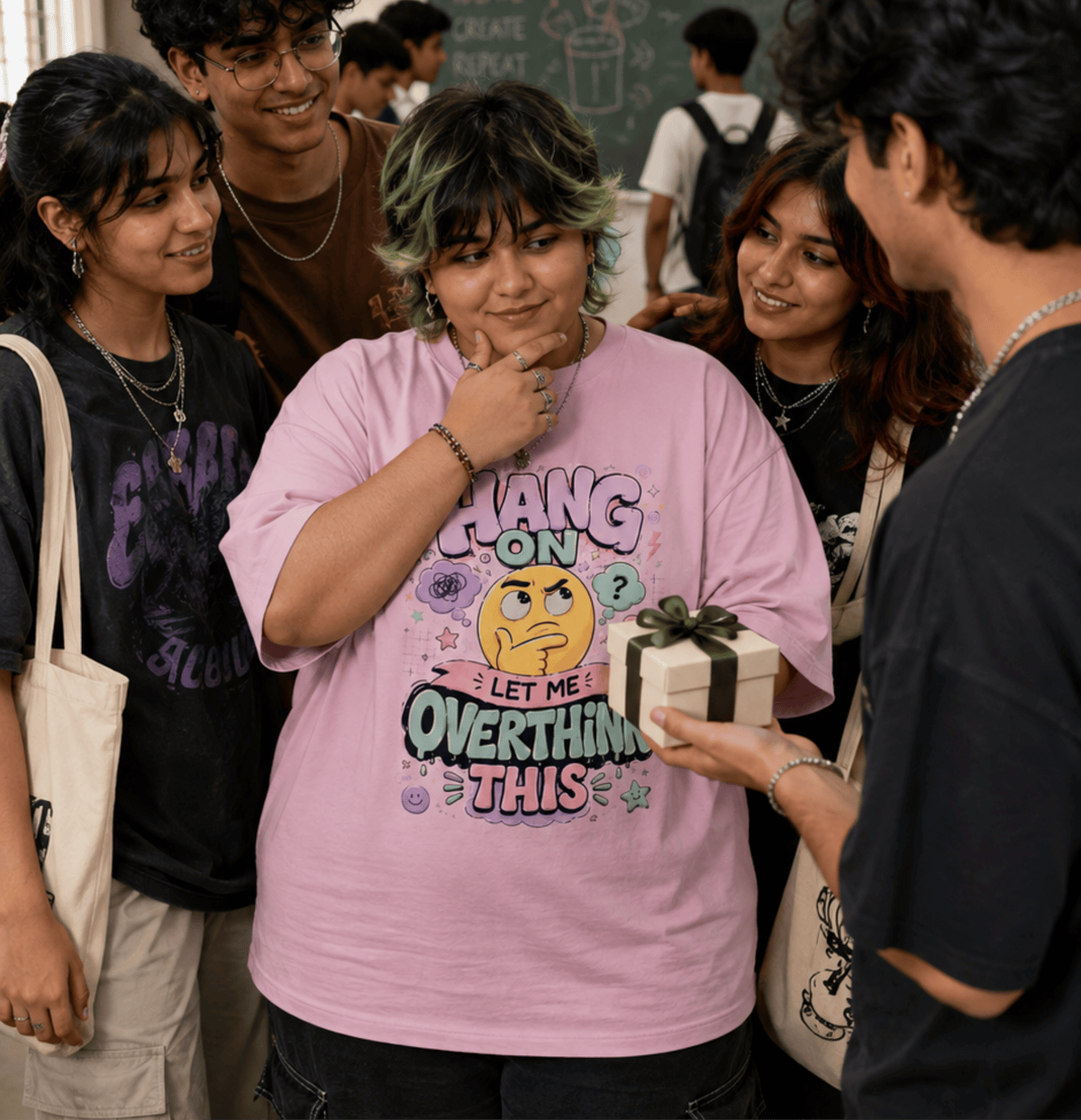 A group of young people in a casual indoor setting, with the central focus on a person wearing a light pink graphic T-shirt that reads 'HANG ON LET ME OVERTHINK THIS' with a yellow face illustration. A hand holds a small gift box near the person.