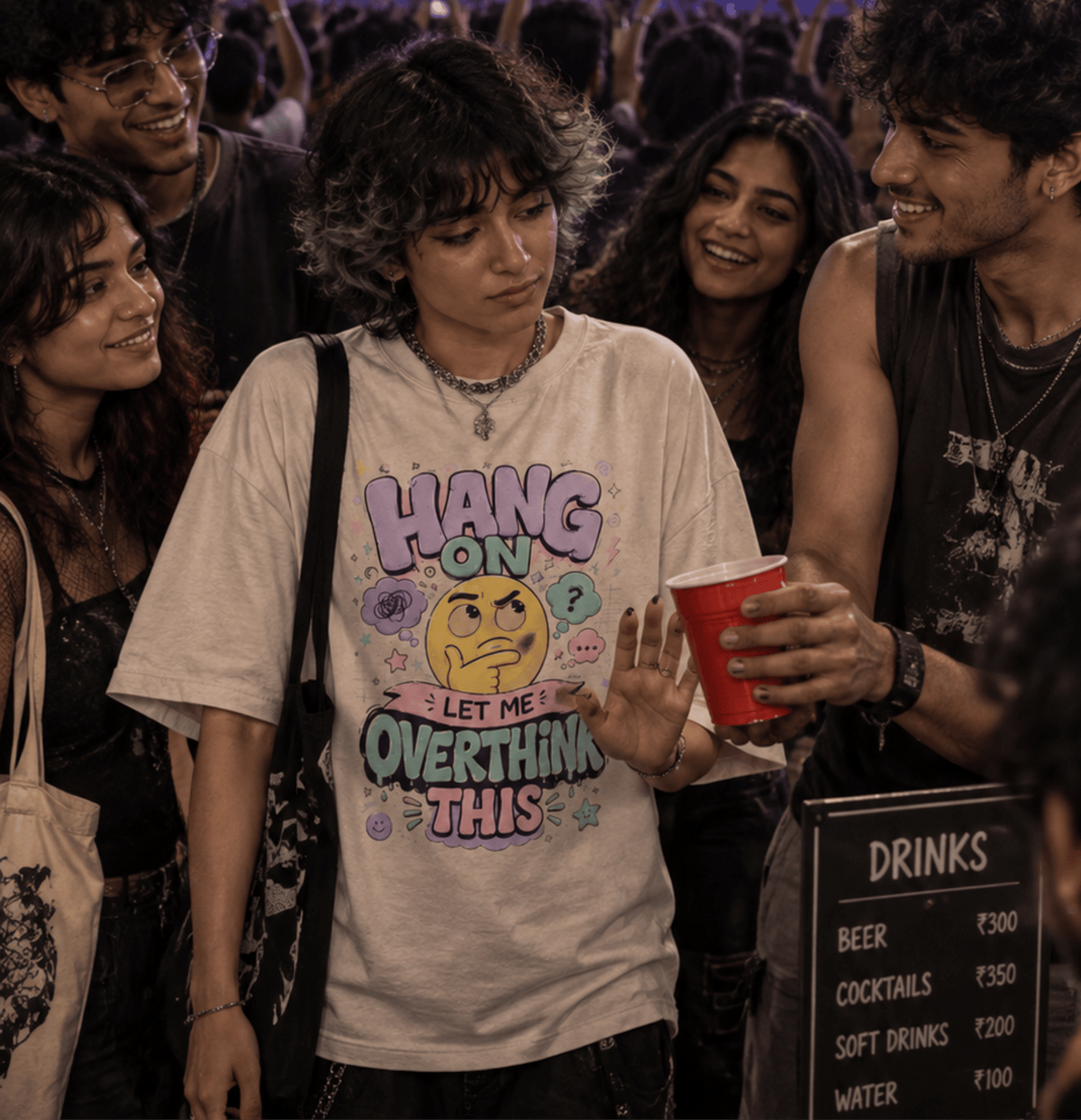 A person standing in a crowded party scene wearing a light beige graphic T-shirt with colorful text reading 'HANG ON LET ME OVERTHINK THIS' and a yellow thinking face graphic. The person holds a red cup and is surrounded by friends at a night event, with a drinks menu sign visible in the foreground.