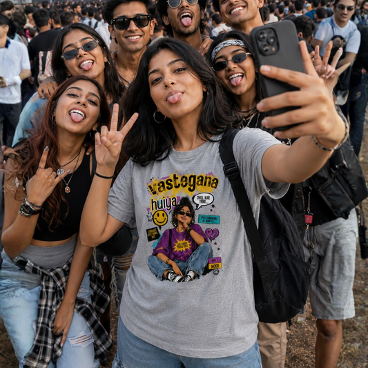A group of young friends taking a selfie outdoors, with the central girl wearing a light gray graphic T-shirt that reads 'Vastegana huiya' with a colorful cartoon-style print. The scene is casual and lively, with other people visible in the background.