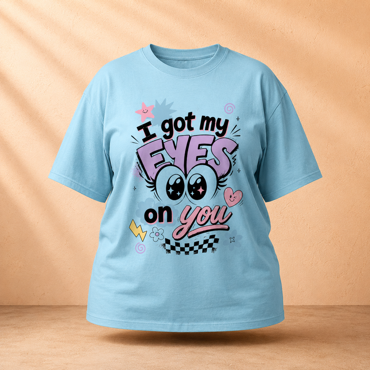 A light blue short-sleeve Oversized Classic T-Shirt with a colorful graphic print that reads 'I got my EYES on you' featuring cartoon eyes, hearts, and checkered details, displayed against a warm beige background with soft shadows.