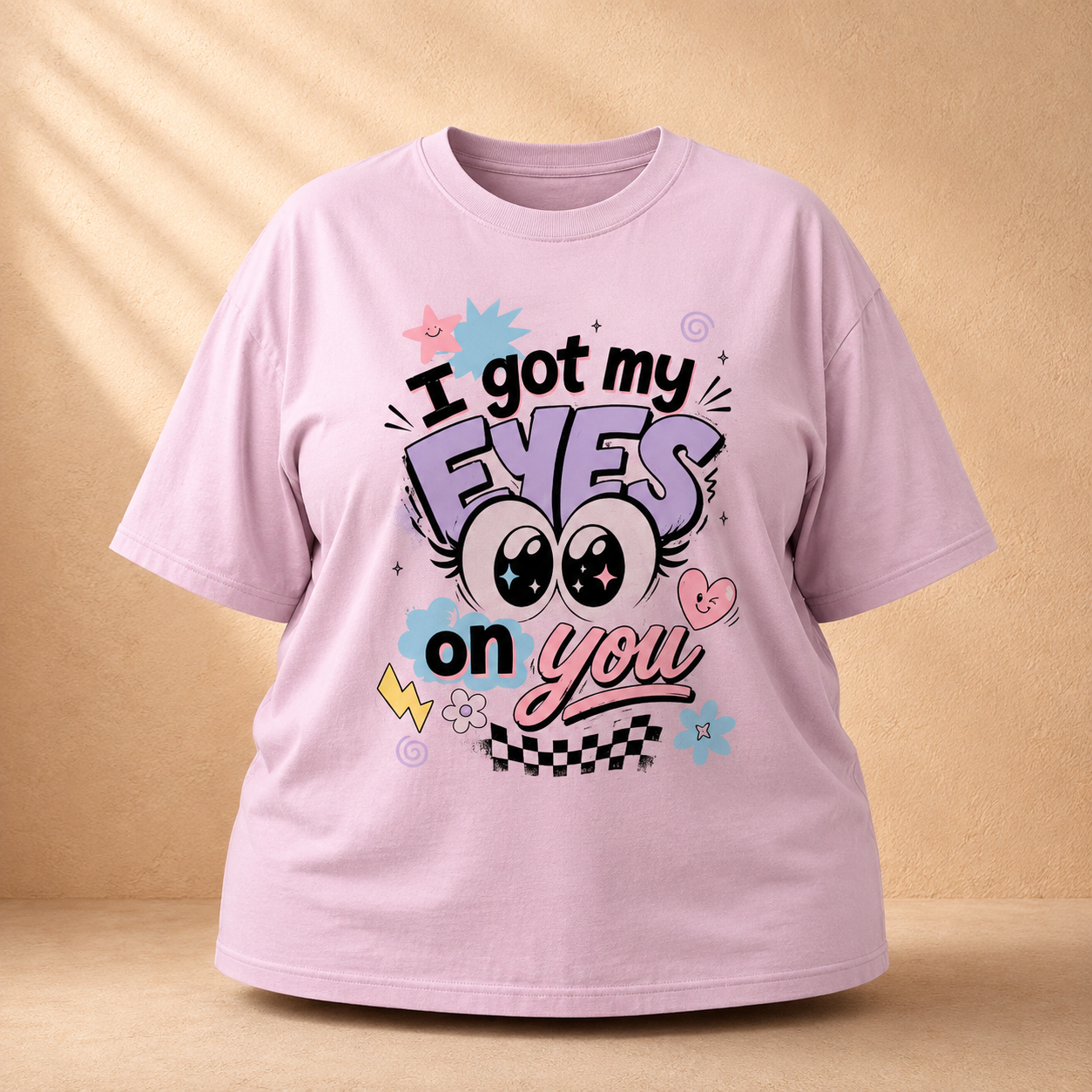 A light pink short-sleeve graphic Oversized Classic T-Shirt displayed against a warm beige background with soft shadow overlays. The graphic features the text 'I got my EYES on you' in stylized black, purple, and pink lettering, accompanied by cartoon eyes, a pink heart, and small decorative doodles.