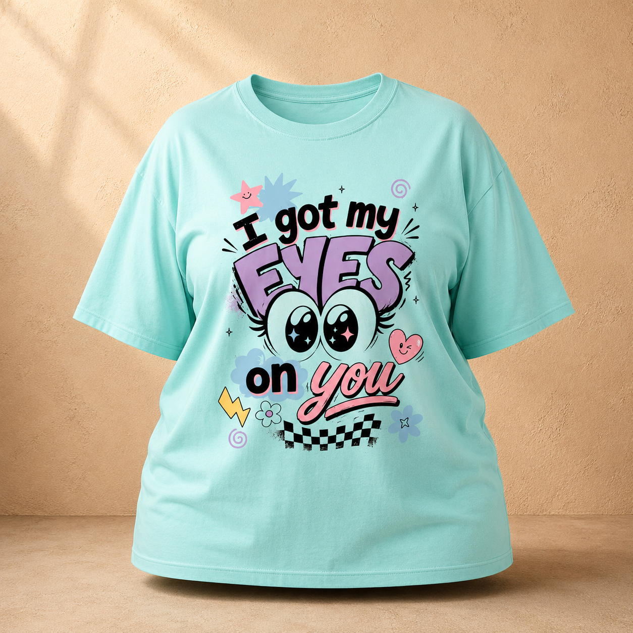 A mint green graphic Oversized Classic T-Shirt displayed against a textured beige wall. The shirt features a colorful retro-style graphic that reads 'I got my EYES on you' with cartoon eyes, stars, and checkers.