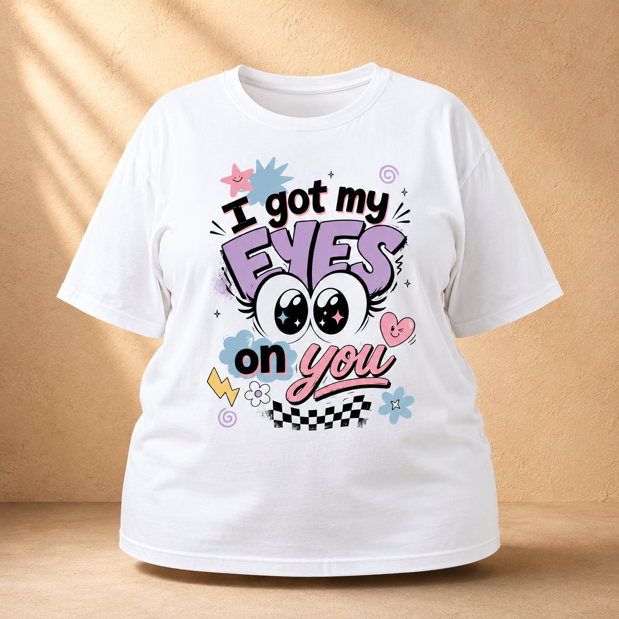 A white short-sleeve graphic Oversized Classic T-Shirt displayed against a warm beige textured background with soft diagonal shadows. The shirt features a colorful cartoon-style graphic with the text 'I got my EYES on you' in black, purple, pink, and blue lettering, accompanied by cartoon eyes, a pink heart, and small decorative accents.