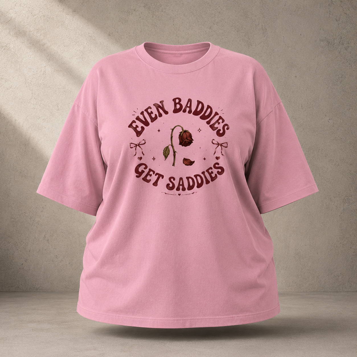 A Flamingo short-sleeve t-shirt with a vintage-style graphic on the chest that reads 'EVEN BADDIES GET SADDIES' around an illustration of a sad rose, shown against a textured gray wall background.