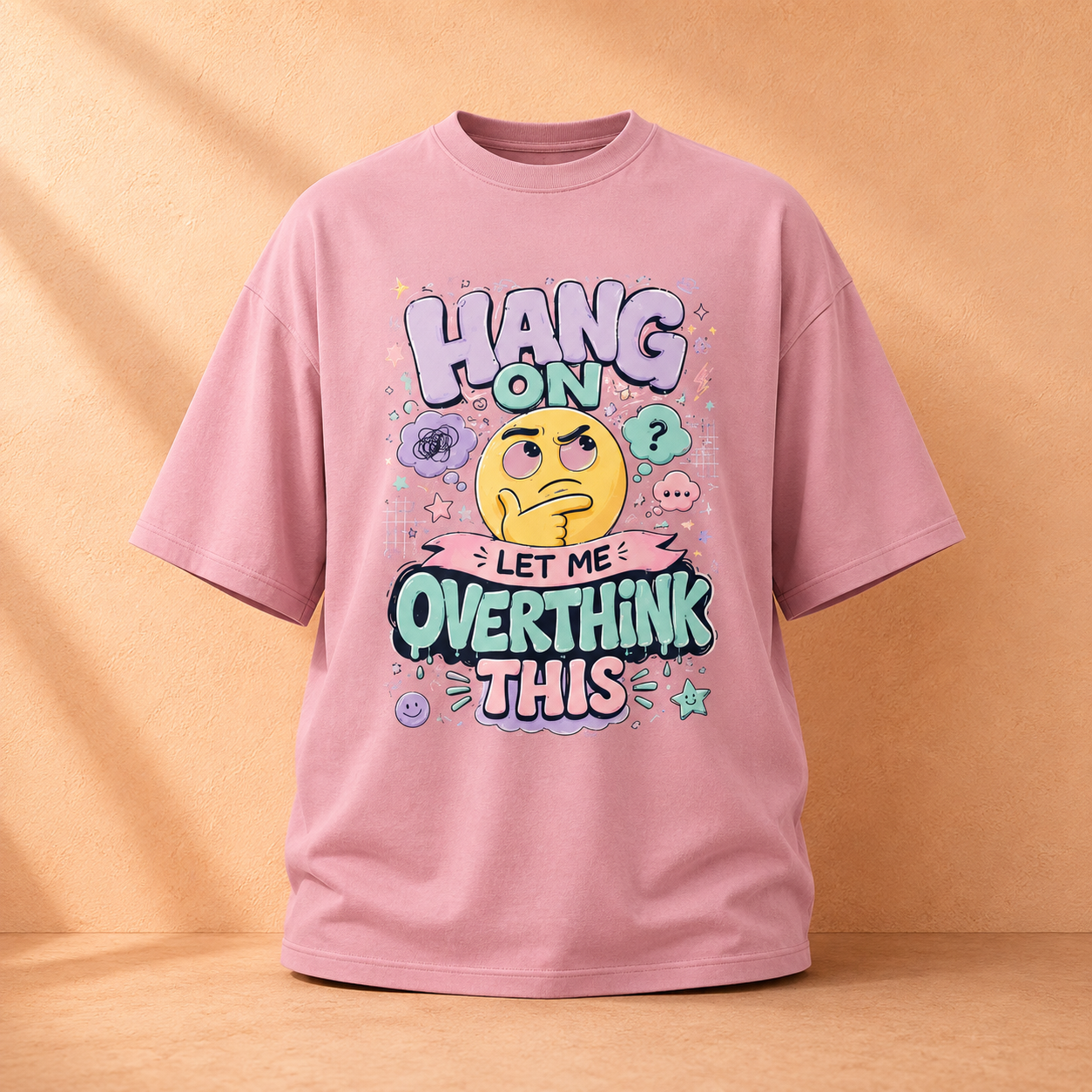 A pink graphic t-shirt displayed against a warm beige textured background. The t-shirt features a colorful cartoon design with a grumpy yellow emoji face and the text 'HANG ON LET ME OVERTHINK THIS'.