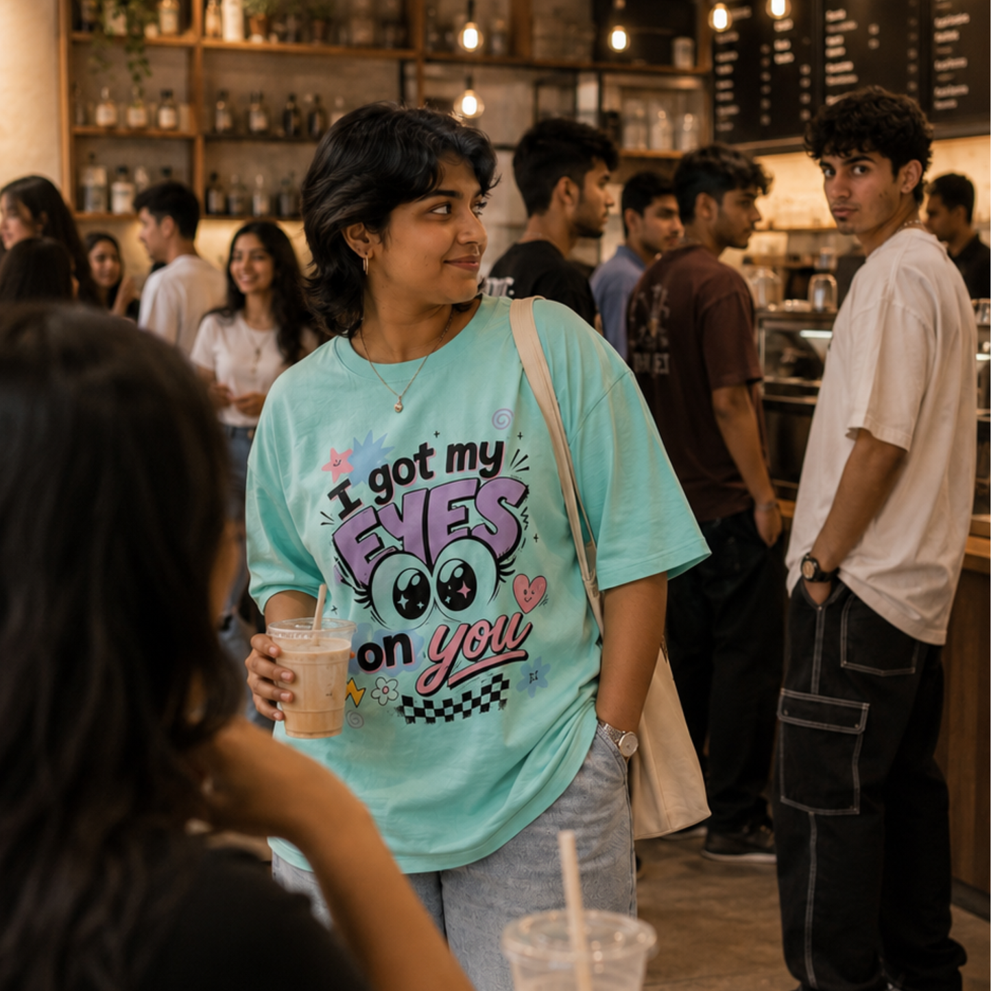 A woman in a busy cafe setting wearing a light blue graphic t-shirt that reads 'I got my EYES on you' with cartoon eyes. She is holding an iced coffee and looking to the side, with other cafe patrons in the background.