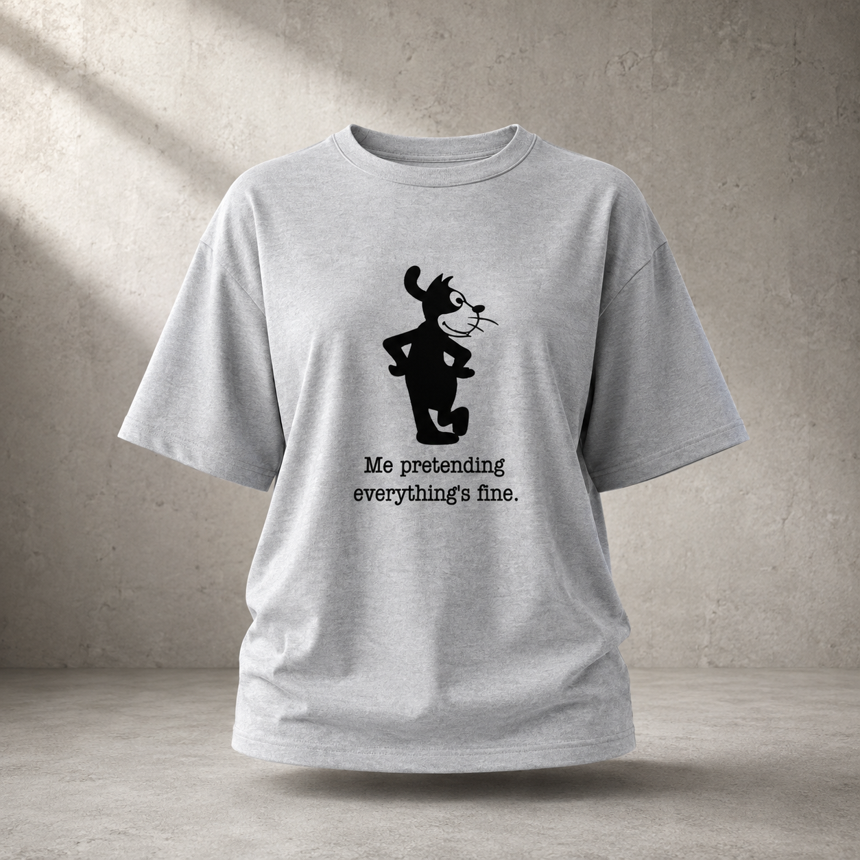 A light heather gray short-sleeve t-shirt with a black graphic of a cartoon cat and the text 'Me pretending everything's fine.' printed on the front, displayed against a textured concrete wall background.
