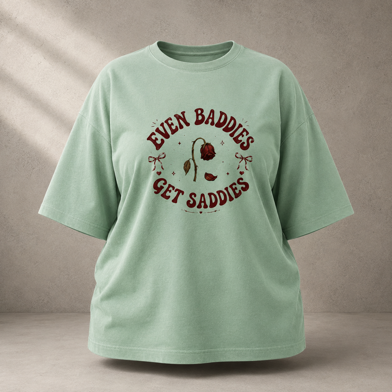 A Jade short-sleeve t-shirt with a dark red graphic print on the front that reads 'EVEN BADDIES GET SADDIES' around an illustration of a rose, displayed against a textured gray wall.
