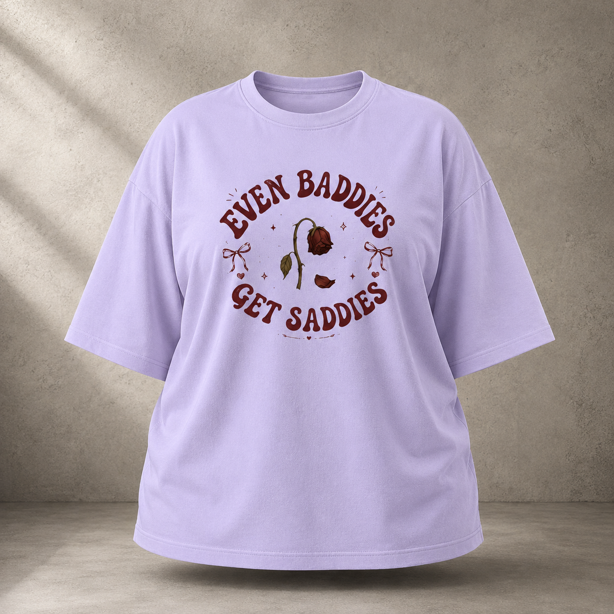 A lavender short-sleeve t-shirt with a dark red graphic print on the front that reads 'EVEN BADDIES GET SADDIES' around a single rose illustration, displayed against a textured gray concrete wall background.