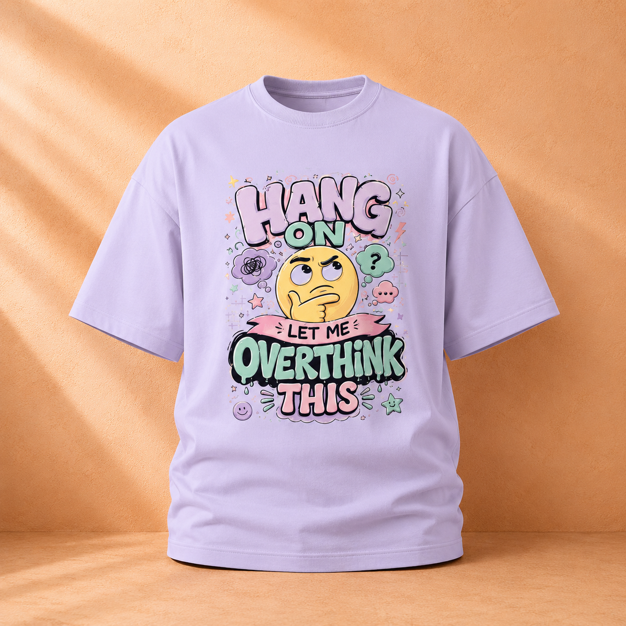 A light lavender short-sleeve t-shirt with a colorful graphic on the front that reads 'HANG ON LET ME OVERTHINK THIS' around a yellow thinking face, displayed against a warm beige background with soft shadows.
