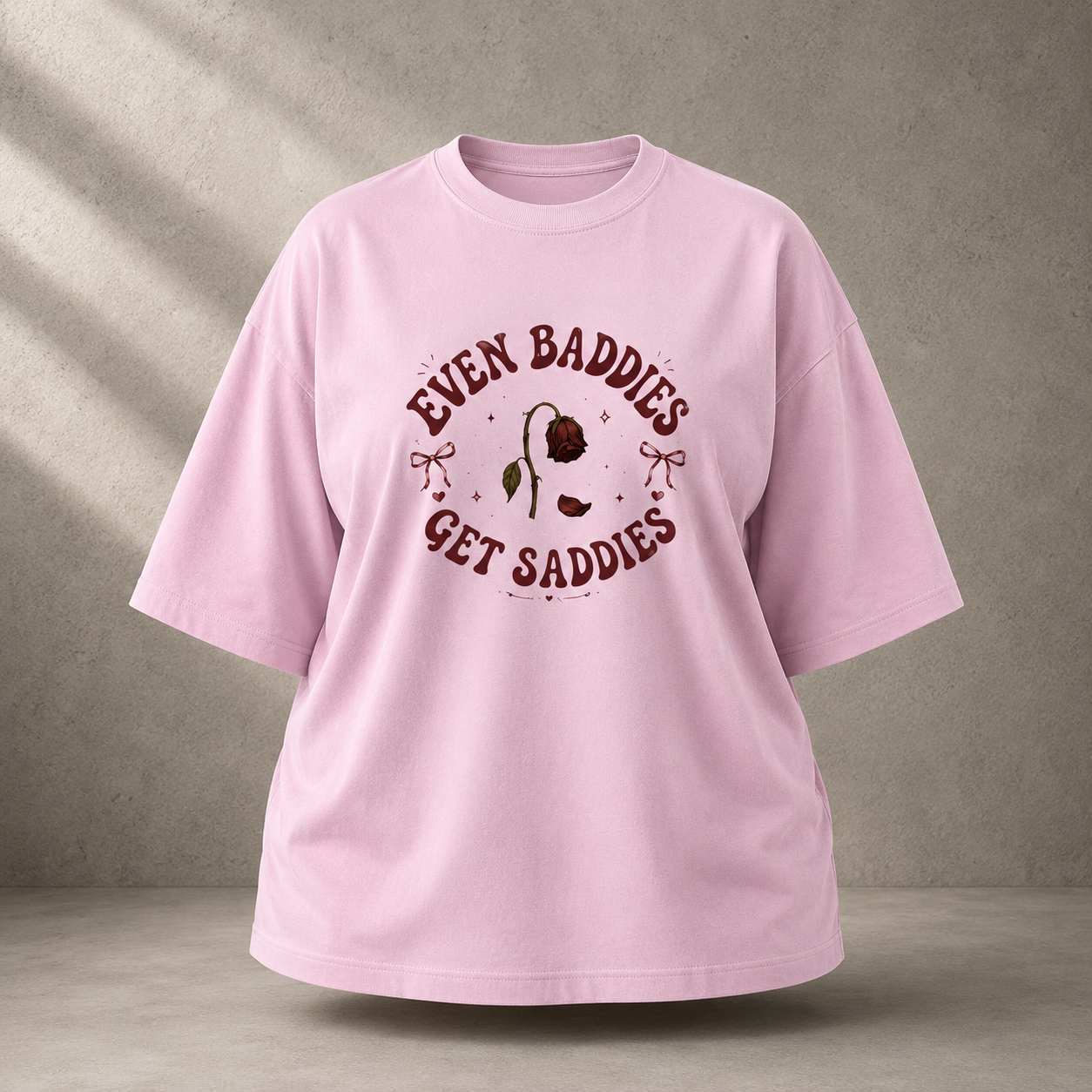 A light baby pink oversized short-sleeve t-shirt with a dark red graphic print on the front that reads 'EVEN BADDIES GET SADDLES' around a rose illustration, displayed against a textured gray wall with soft diagonal shadows.
