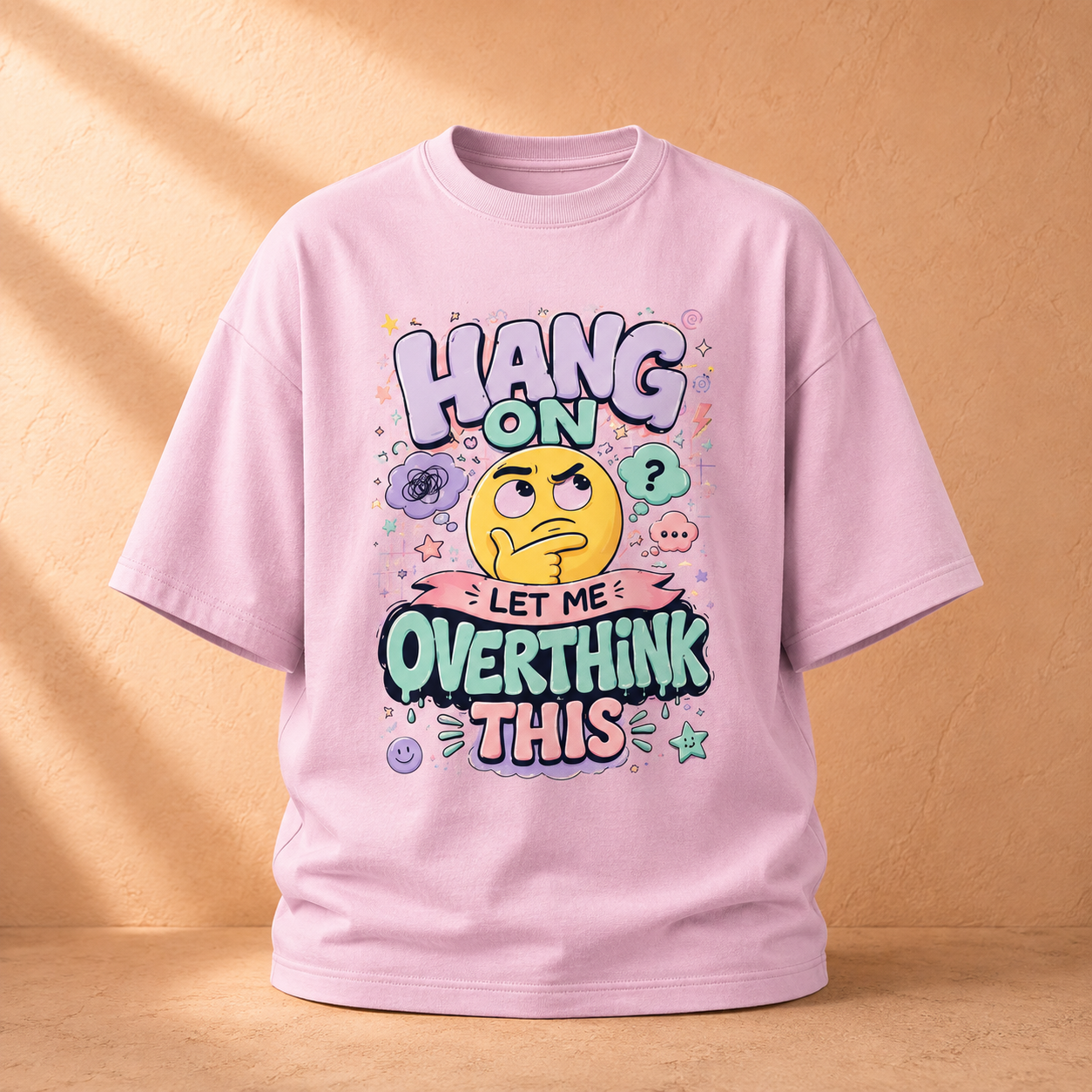 A light pink short-sleeve t-shirt with a colorful graphic print on the front, displayed against a warm beige textured background with soft diagonal shadows.