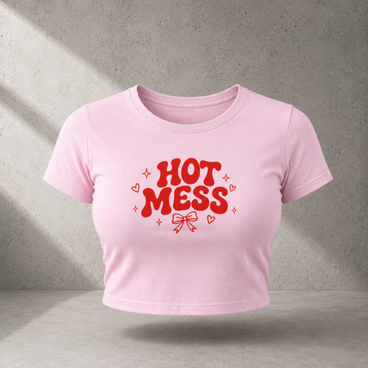 A light pink cropped short-sleeve t-shirt with a red graphic text reading 'HOT MESS' and small heart and sparkle accents, shown against a textured gray wall with soft diagonal shadows.