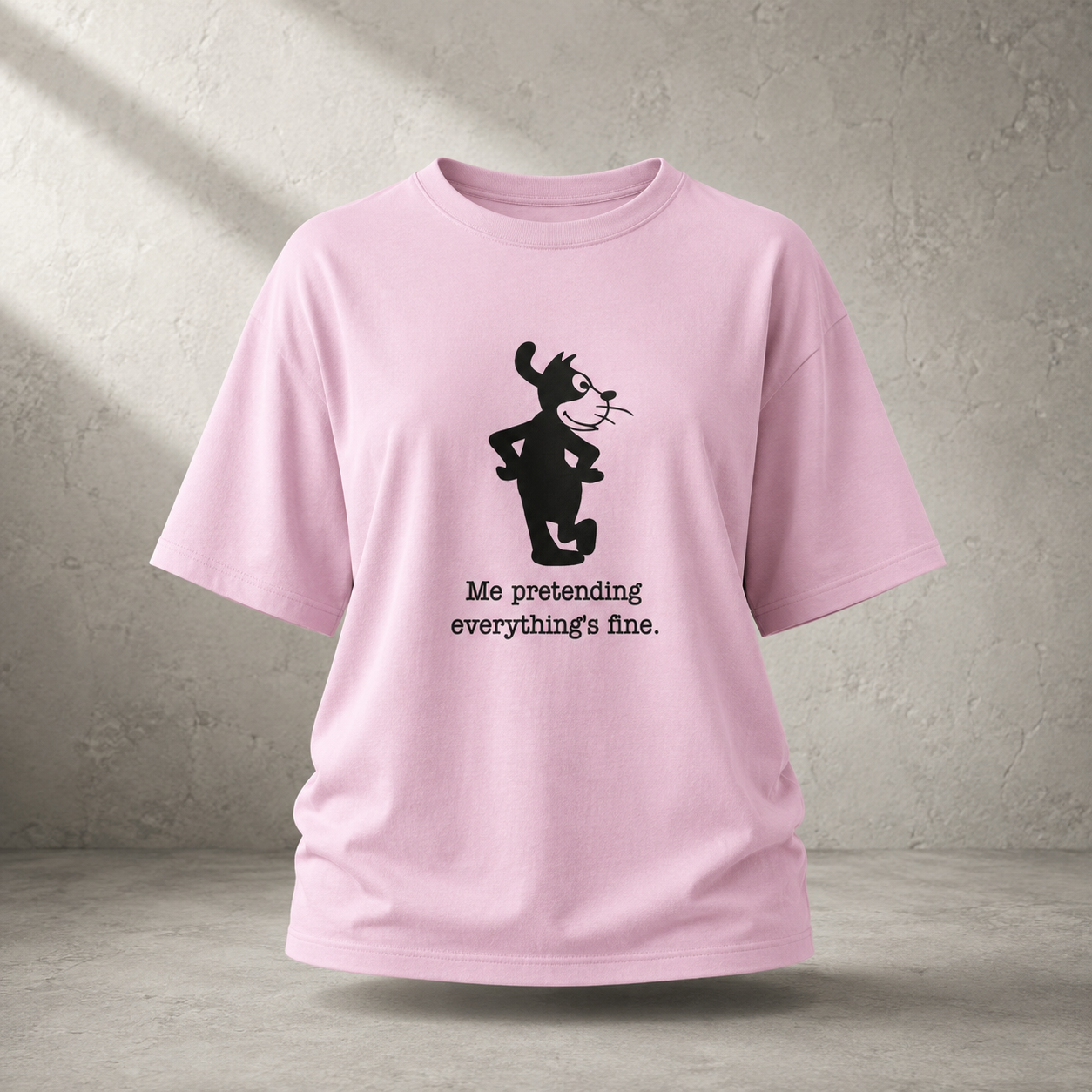 A light pink short-sleeve t-shirt with a black graphic of a cartoon cat and the text 'Me pretending everything's fine.' printed on the front, displayed against a textured gray concrete wall background.
