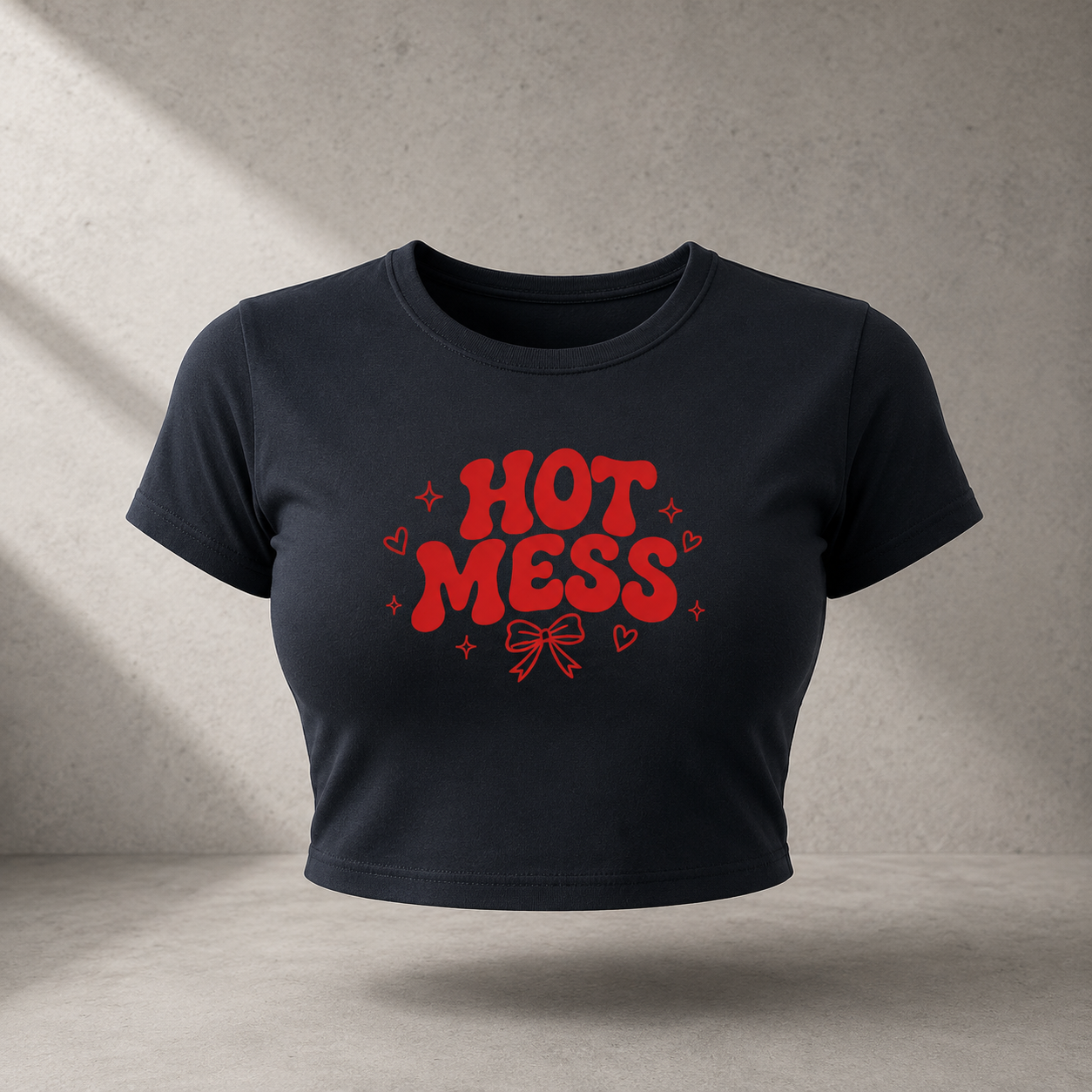 A cropped black t-shirt with a red graphic print reading 'HOT MESS' in a bold retro font, surrounded by small hearts and stars, displayed against a textured gray concrete wall with diagonal shadows.