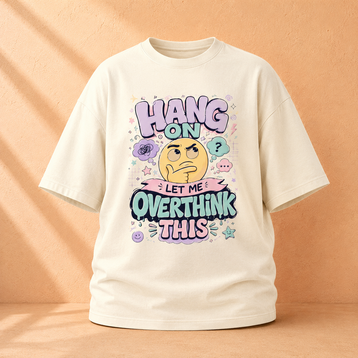 An off-white short-sleeve t-shirt with a colorful graphic print that reads 'HANG ON LET ME OVERTHINK THIS' around a thinking emoji, displayed against a warm peach background with soft shadow overlays.