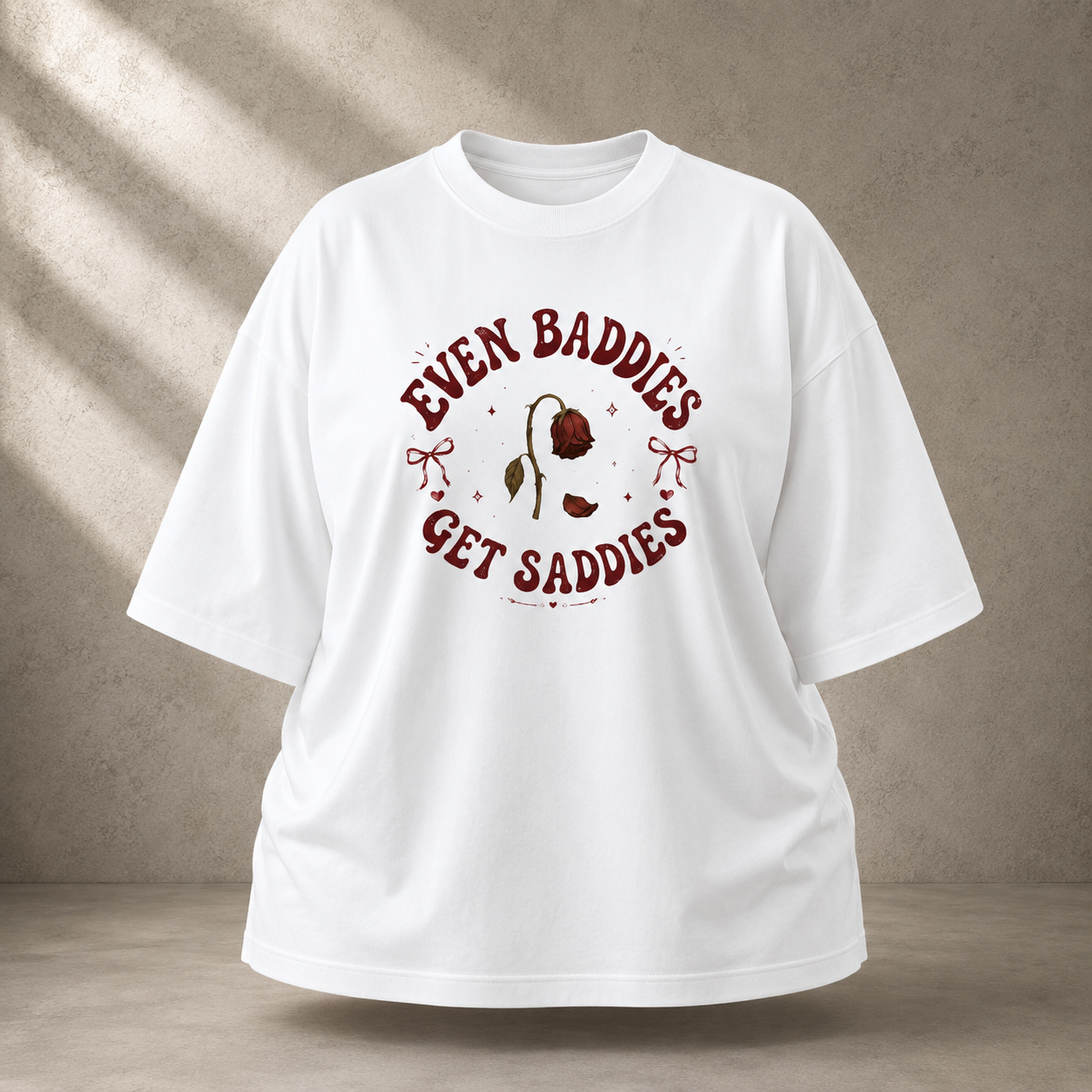 A white oversized short-sleeve t-shirt with a vintage-style graphic on the chest that reads 'EVEN BADDIES GET SADDIES' in dark red, surrounding a small illustration of a withered rose. The t-shirt is displayed against a textured gray wall with diagonal shadow patterns.