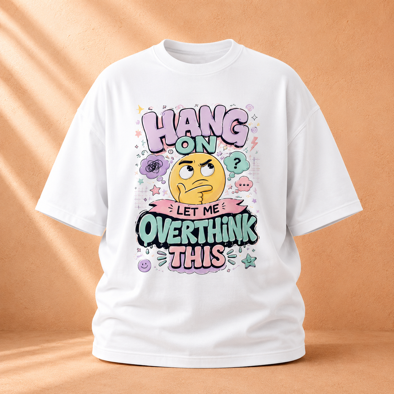 A white short-sleeve graphic t-shirt displayed on a warm, textured peach background with soft diagonal shadows. The graphic features a cartoon face with a thinking expression surrounded by colorful text that reads 'HANG ON LET ME OVERTHINK THIS' in playful pink, purple, and green lettering.