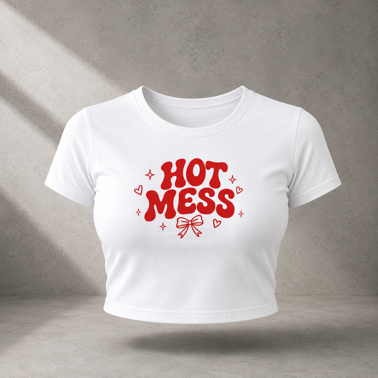 A white short-sleeve cropped T-shirt with a red graphic reading 'HOT MESS' in bold bubble letters, surrounded by small hearts and sparkles, centered on a gray textured studio background.